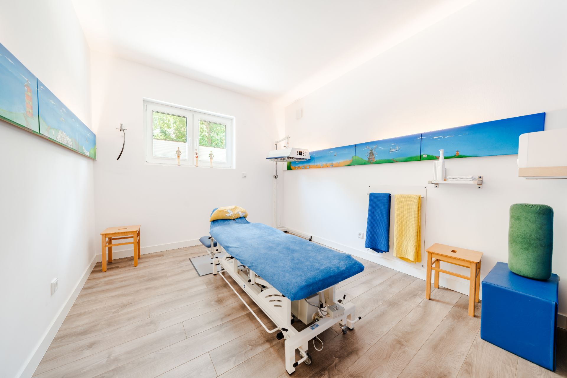 Heller Raum mit einer blauen Behandlungsliege, zwei Fenstern, Stühlen und bunten Handtüchern an der Wand.