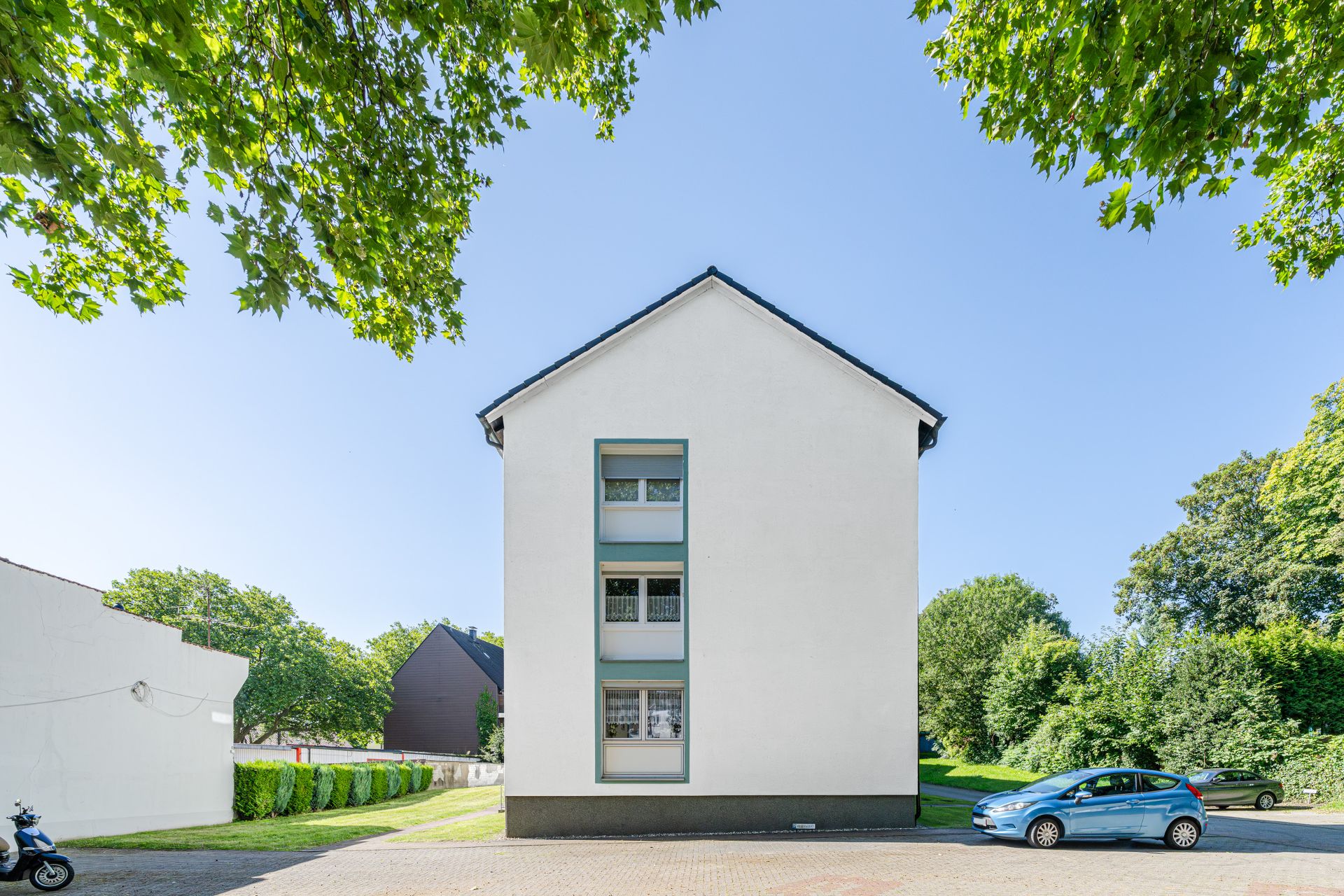 Weiße Hausfassade mit drei Fenstern, davor parkt ein blaues Auto, umgeben von Bäumen.