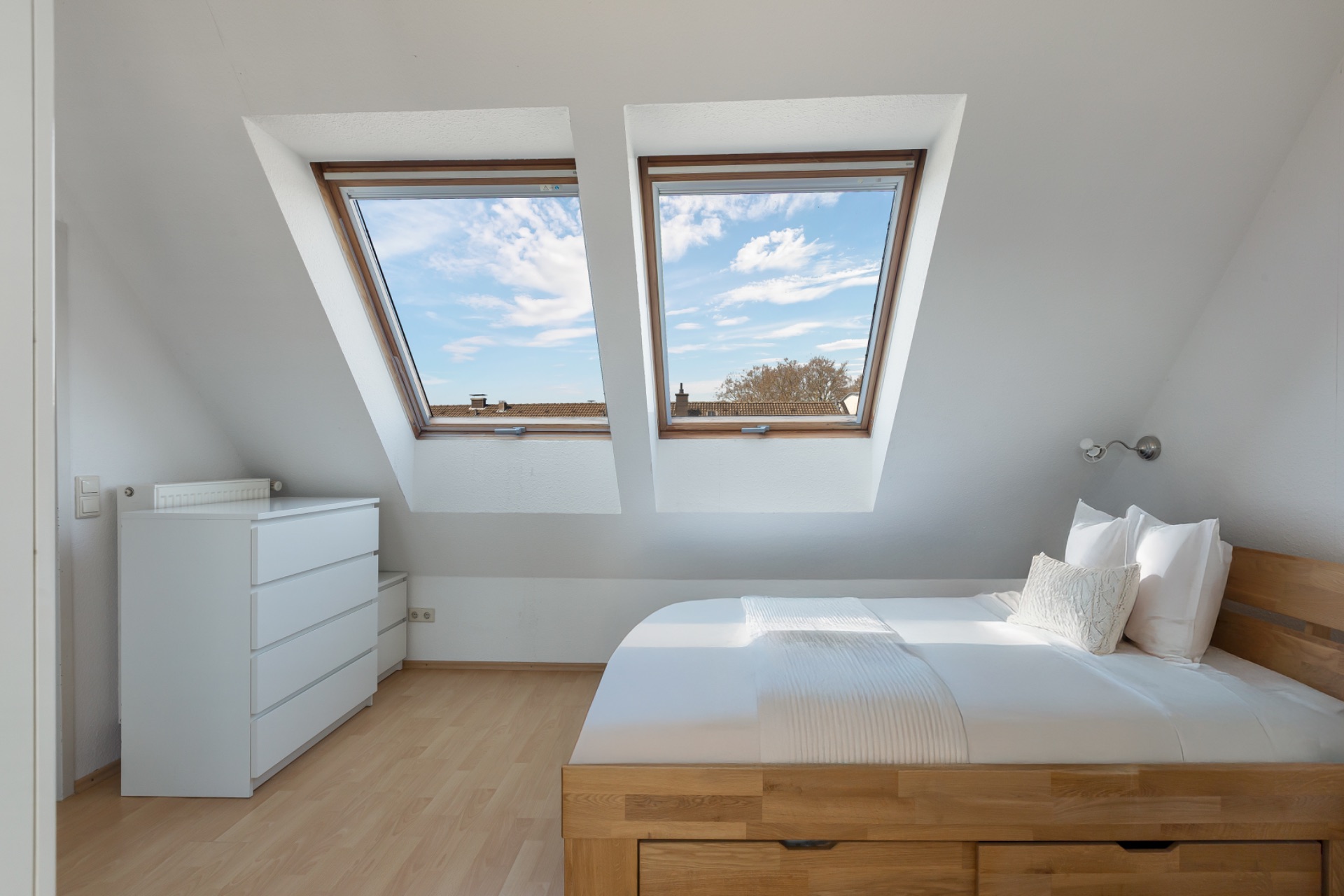 Helles Schlafzimmer mit Holzbett, Kommode und zwei großen Dachfenstern.