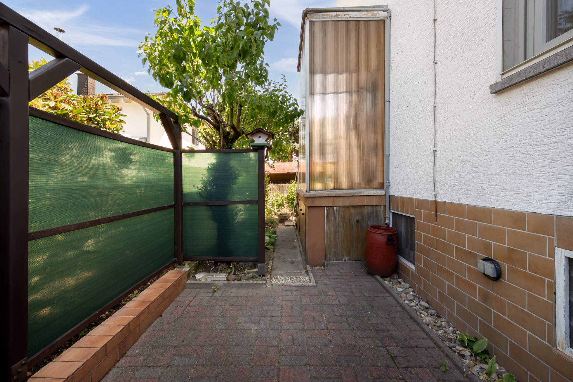 Gepflasterter Weg neben einem Haus mit grünem Zaun, Baum und rotem Behälter.