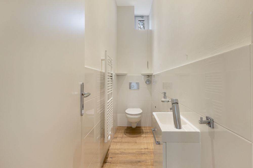 Helles, schmales Badezimmer mit Wandtoilette, kleinem Waschbecken und Holzfußboden.