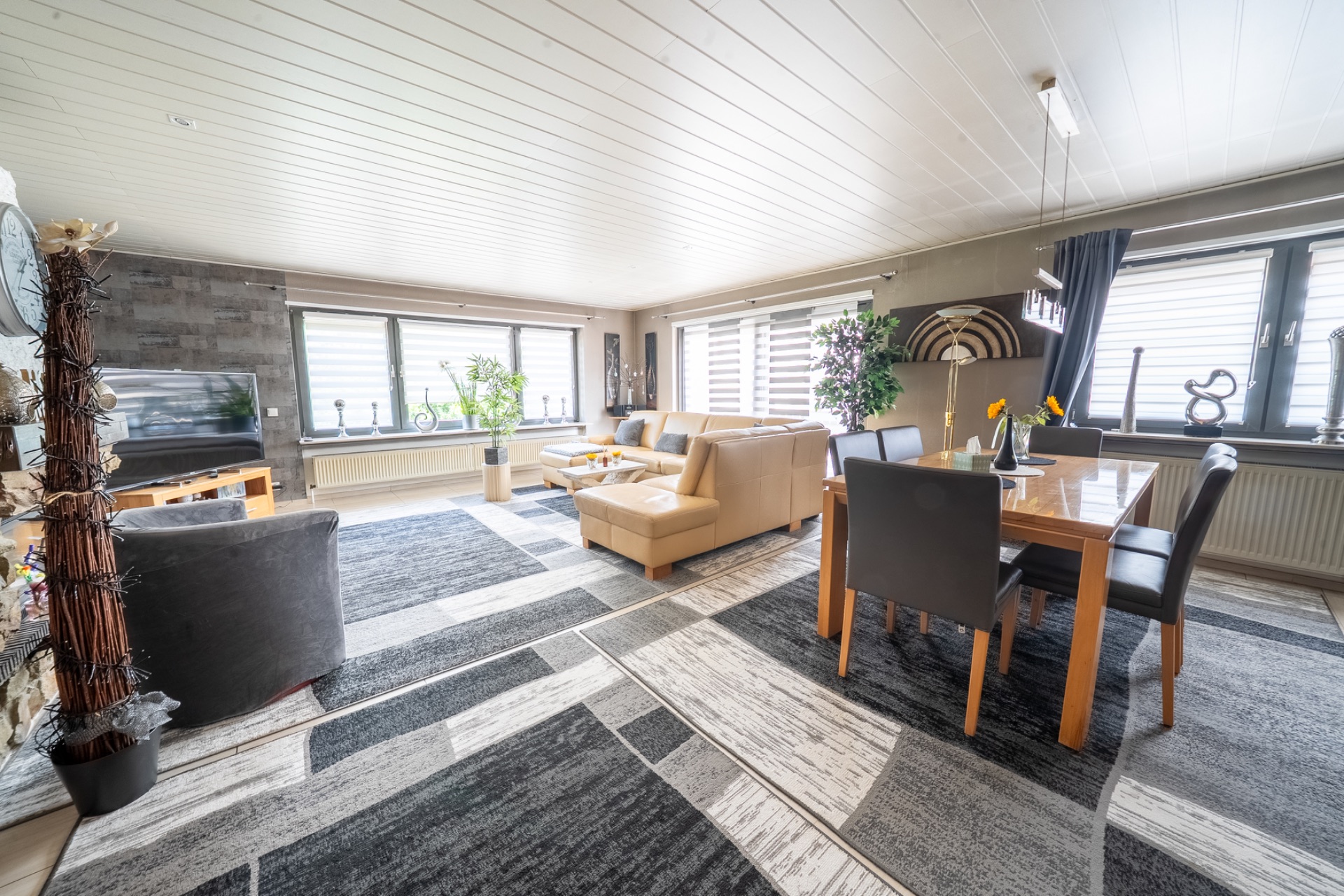 Helles Wohnzimmer mit großen Fenstern, Ledersofa, Esstisch und Teppichboden.
