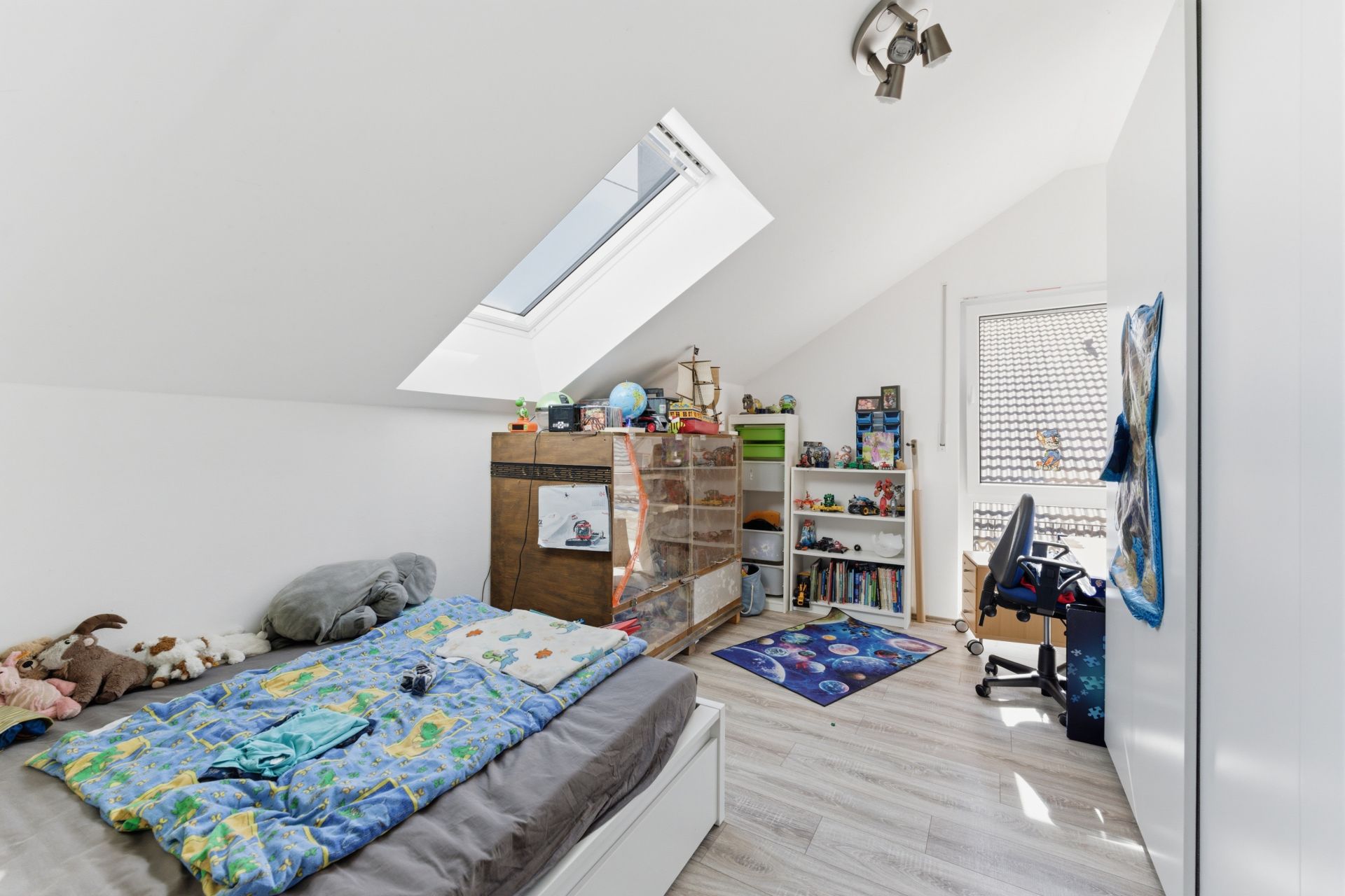 Helles Kinderzimmer mit Bett, Schreibtisch, Regal und Dachfenster unter Schrägdach.