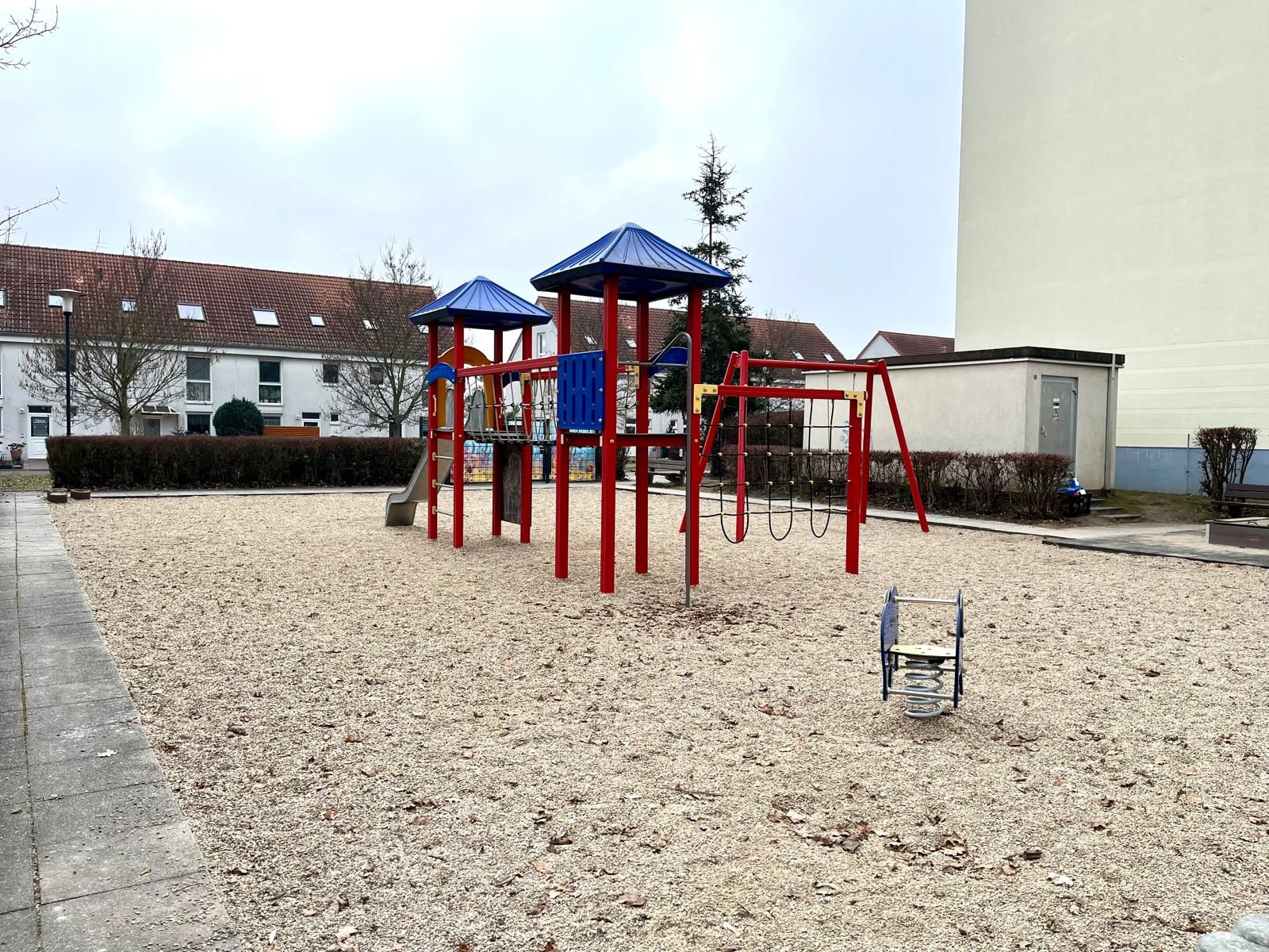 Spielplatz mit Klettergerüst, Rutsche und Schaukeln auf Kiesfläche in einem Wohngebiet.