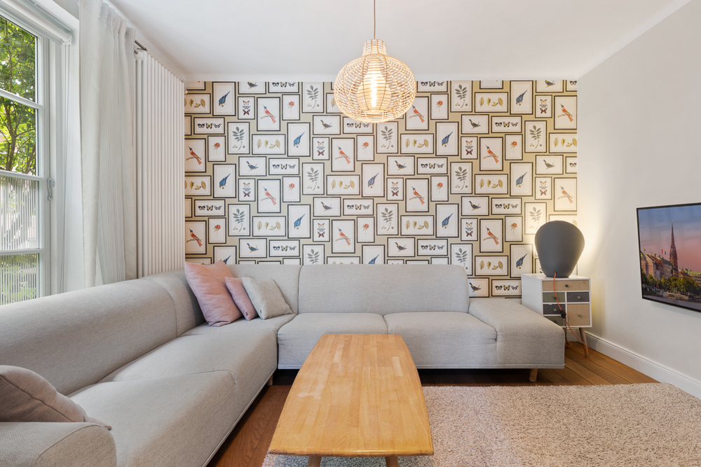 Helles Wohnzimmer mit grauem Ecksofa, Holztisch und Tapete mit Vogelmotiven.