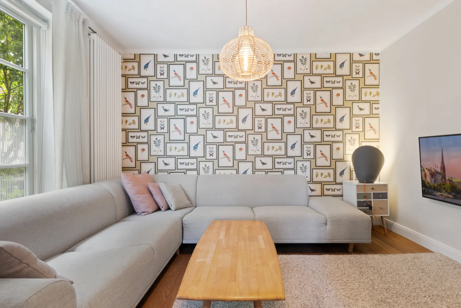 Helles Wohnzimmer mit grauem Ecksofa, Holztisch und Tapete mit Vogelmotiven.