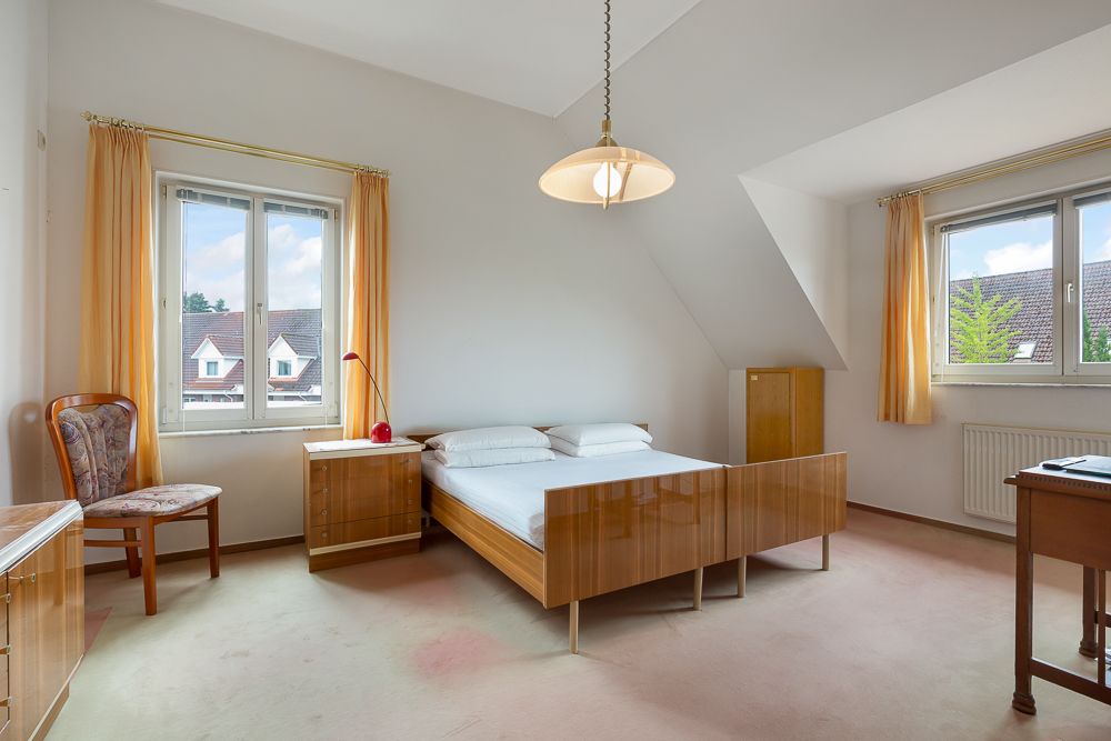 Helles Schlafzimmer mit Doppelbett, zwei Fenstern, Holzmöbeln und gelben Vorhängen.