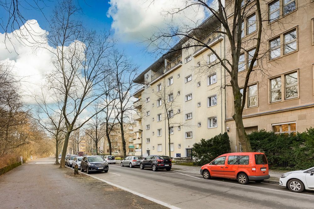 Wohngebäude mit mehreren Etagen an einer Straße mit parkenden Autos und Bäumen.