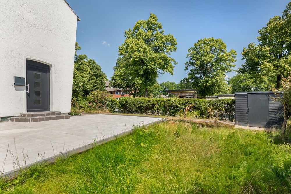 Eingangsbereich eines Hauses mit Terrasse, Rasen und Gartenhaus bei sonnigem Wetter.