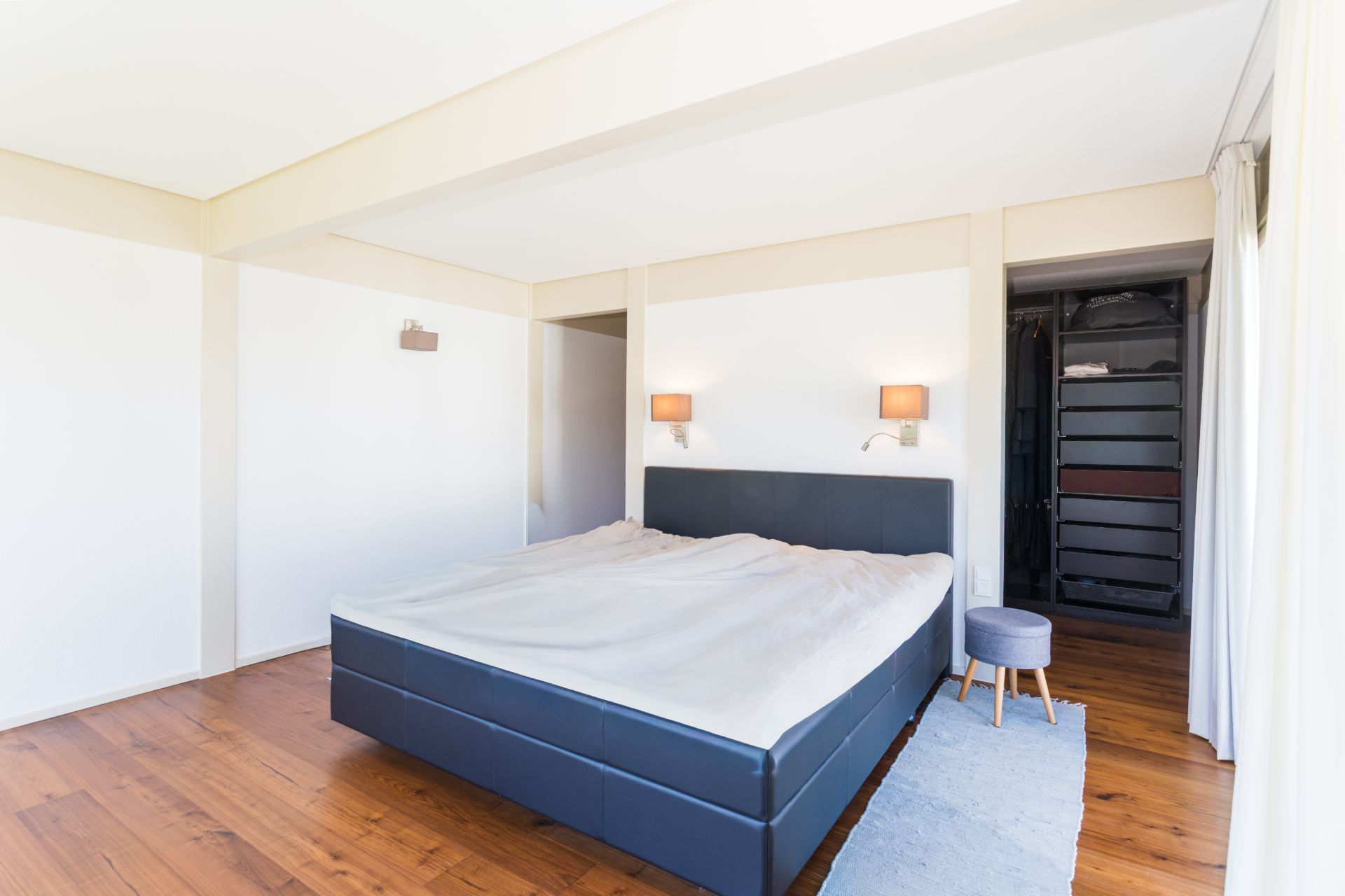 Modernes Schlafzimmer mit großem Bett, Teppich, Hocker und Blick in einen offenen Kleiderschrank.