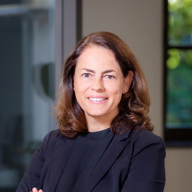 Frau mit braunen Haaren im schwarzen Blazer steht mit verschränkten Armen in einem modernen Raum.