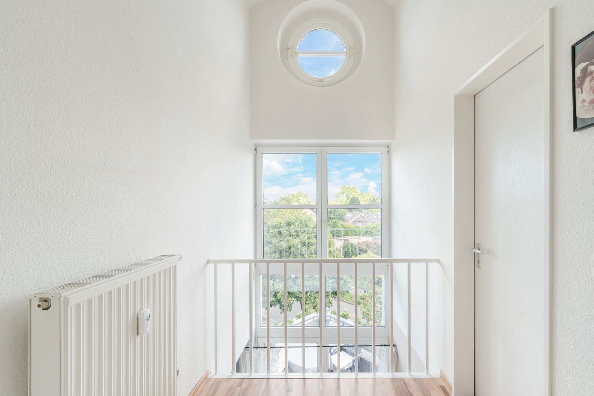 Heller Flur mit großem Fenster, Heizkörper und Tür, Holzfußboden, Blick ins Grüne.