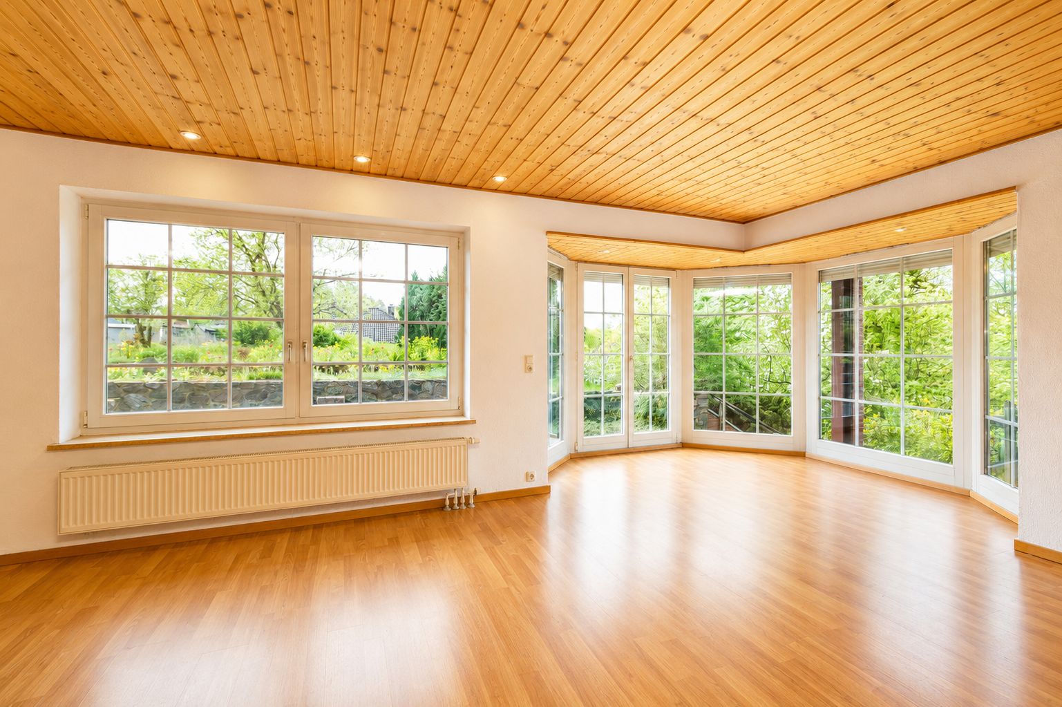 Helles Wohnzimmer mit großen Fenstern, Holzdecke und Parkettboden, Blick ins Grüne.