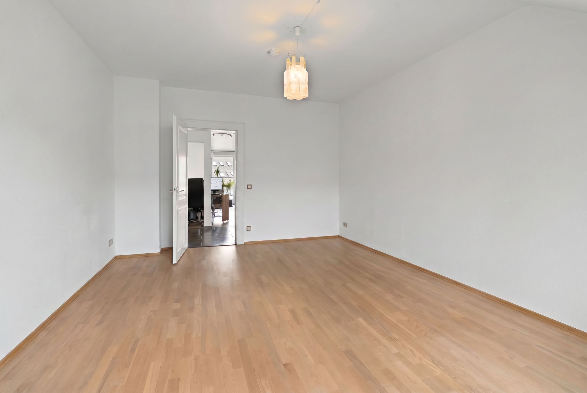 Leerer Raum mit Holzfußboden, weißen Wänden und Deckenlampe, Tür führt in anderen Raum.
