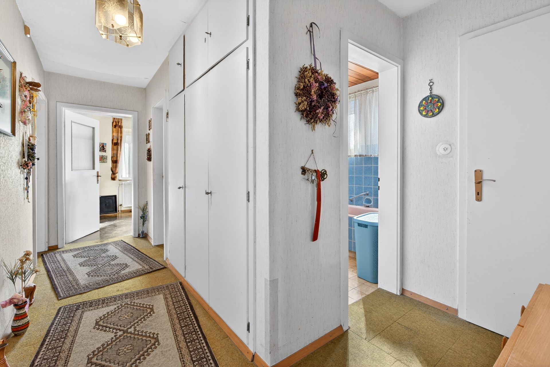 Heller Flur mit weißen Einbauschränken und Teppichen, rechts Zugang zu einem Badezimmer mit blauen Fliesen.