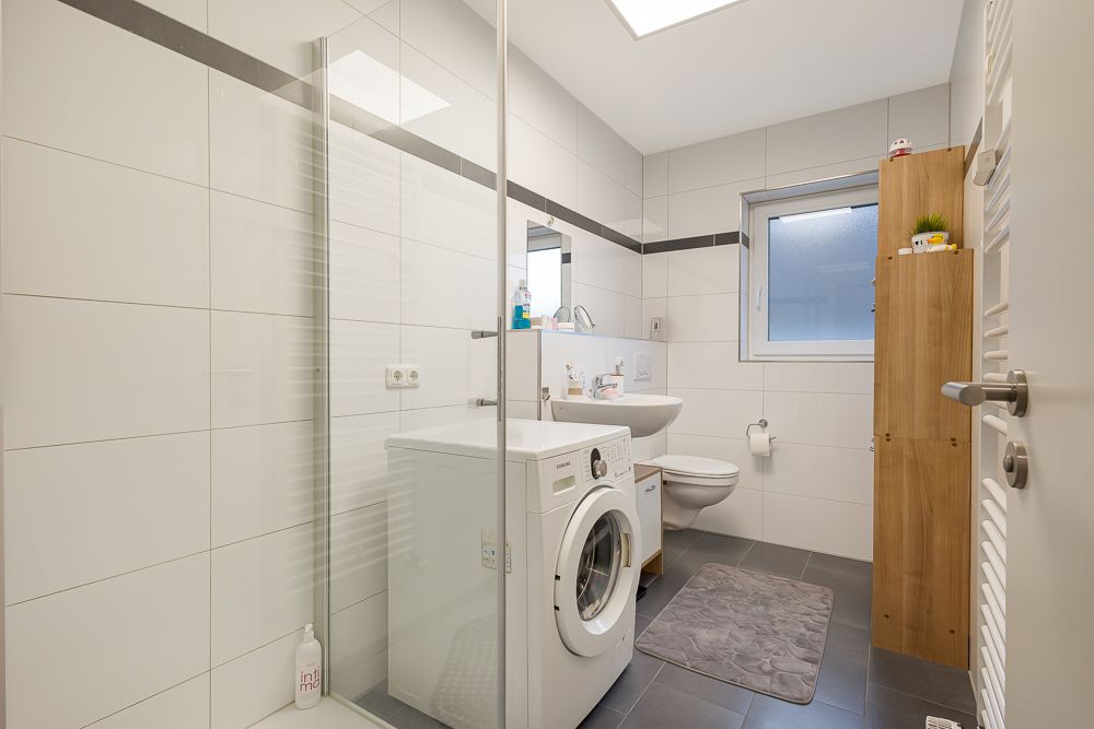 Modernes Badezimmer mit Waschmaschine, Dusche, WC, Waschbecken und Holzregal.