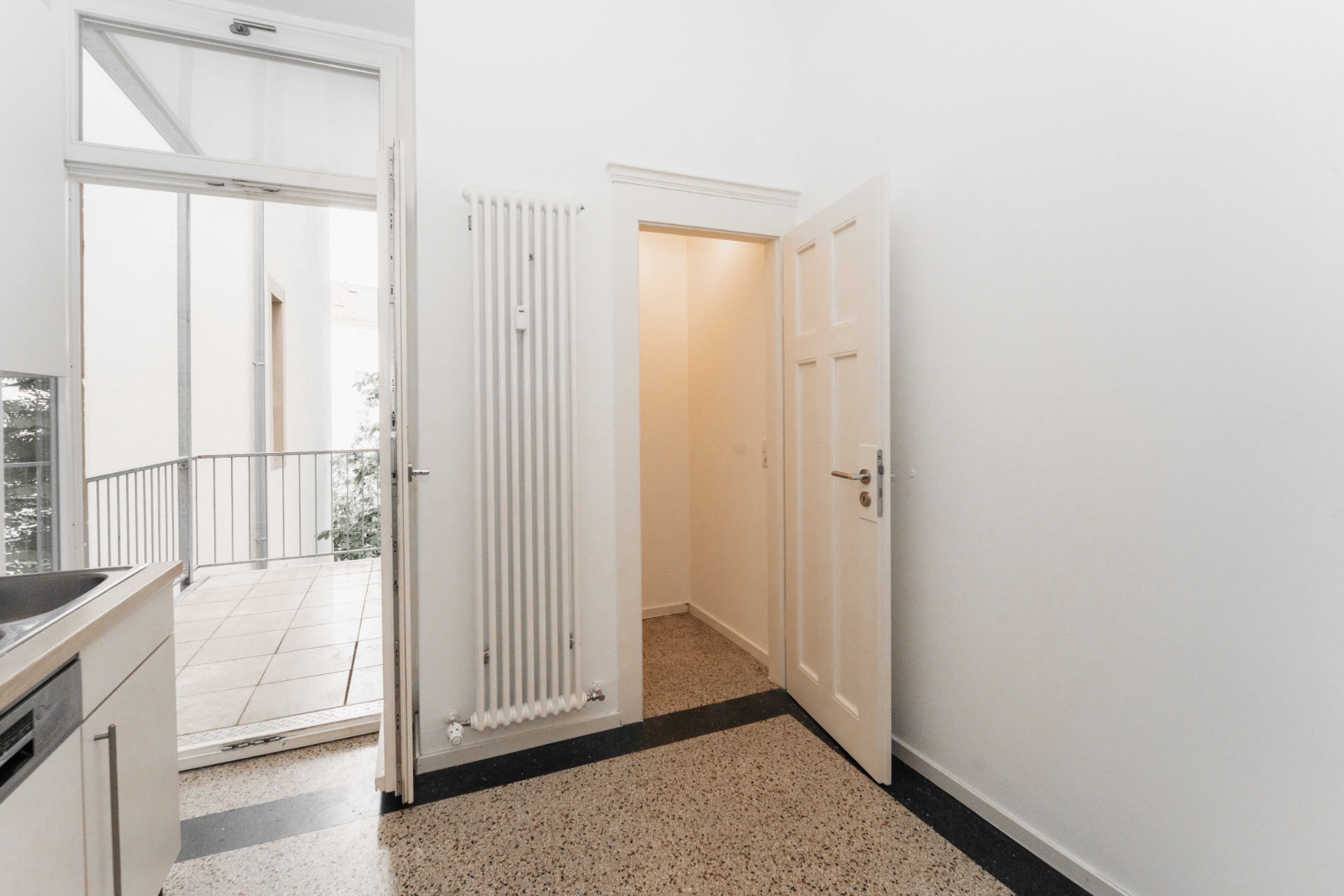 Heller Raum mit offenem Zugang zum Balkon, Heizkörper und geöffneter Tür zu kleinem Flur.