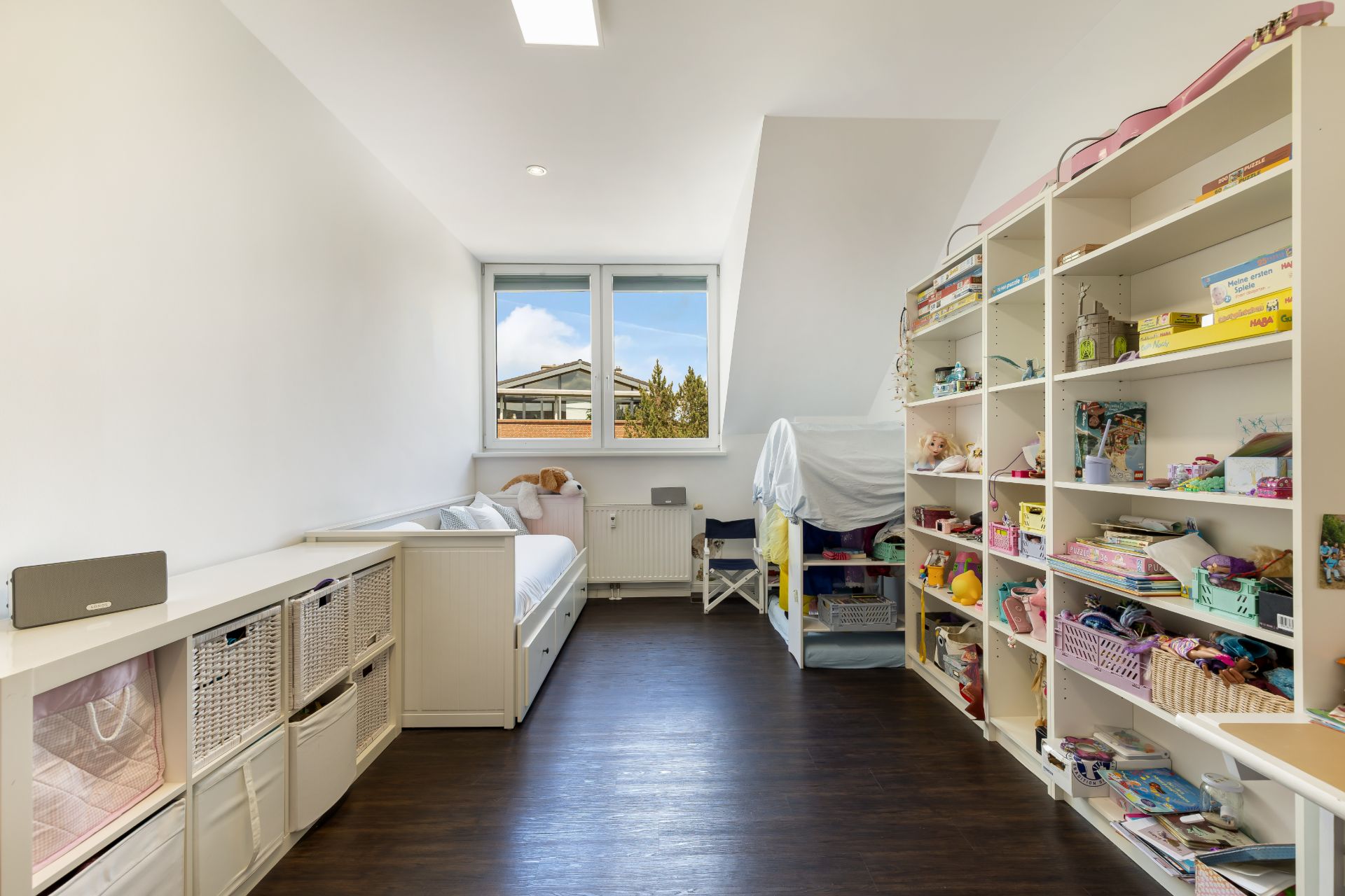 Helles Kinderzimmer mit Regal voller Spielzeug, Einzelbett und Fenster mit Ausblick.