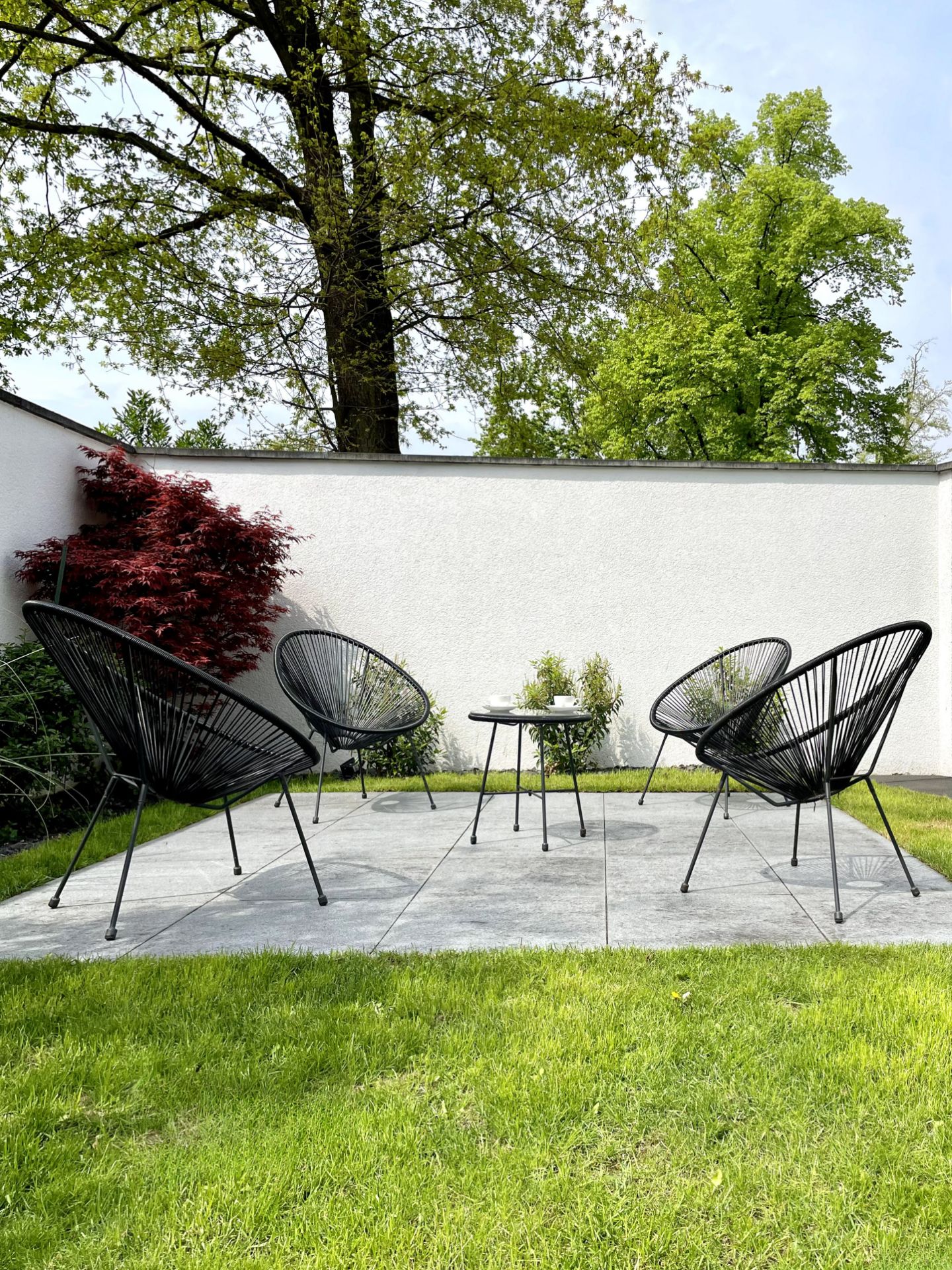 Vier schwarze Gartenstühle und ein kleiner Tisch auf einer gepflasterten Terrasse im Garten.