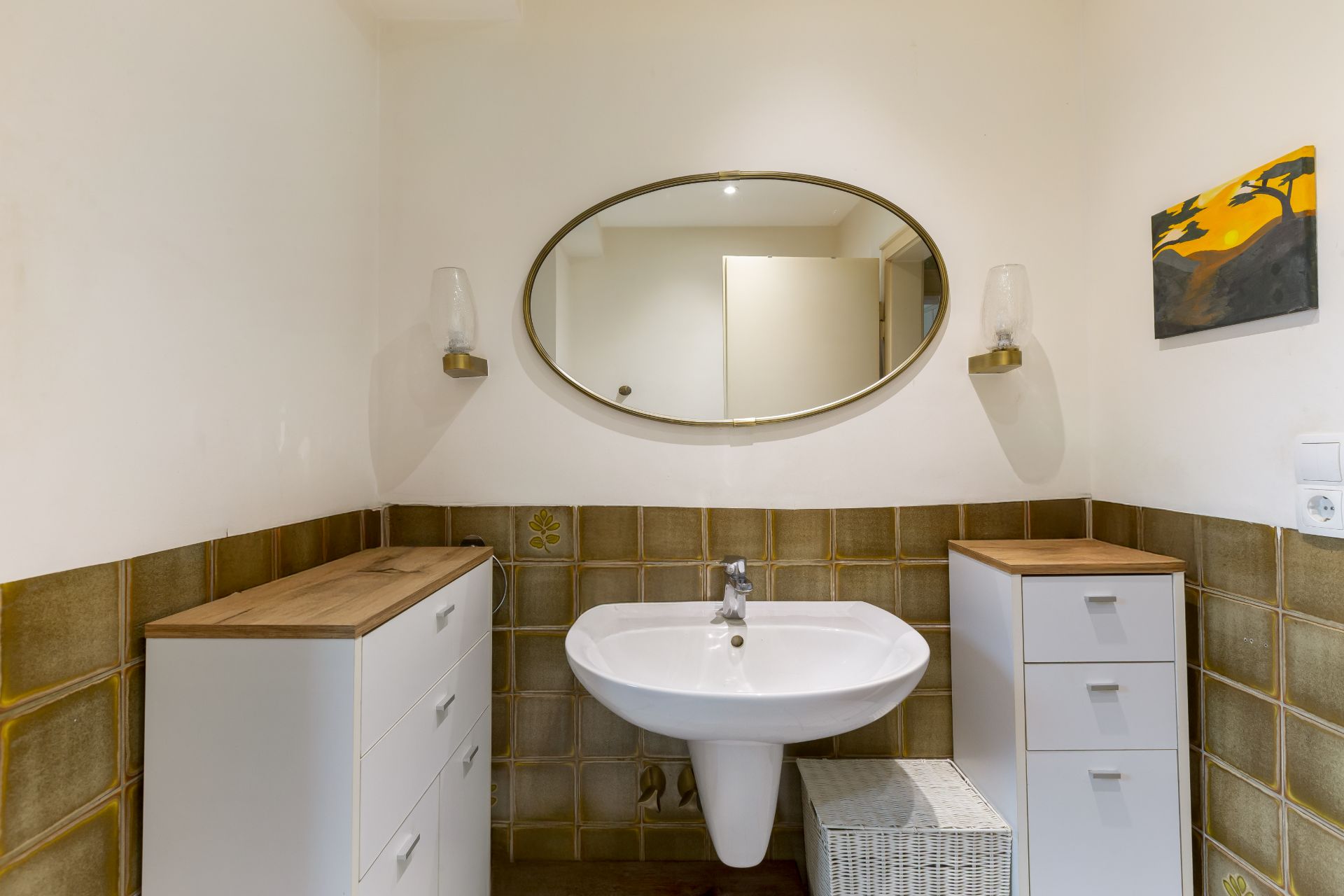 Badezimmer mit Waschbecken, ovalem Spiegel und zwei weißen Schränken.