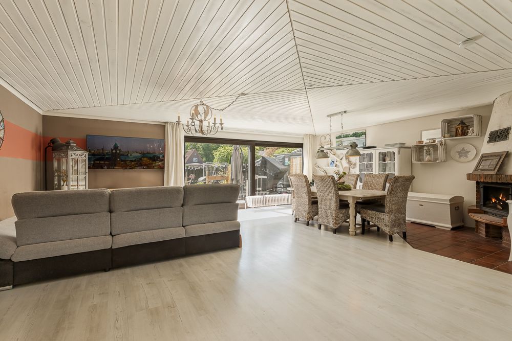 Helles Wohnzimmer mit grauer Couch, Esstisch, Kamin und großem Fenster zur Terrasse.