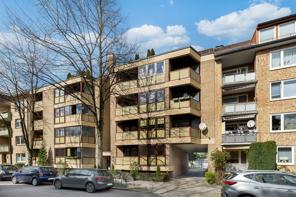 Mehrfamilienhaus mit mehreren Balkonen, geparkten Autos und Bäumen an der Straße.