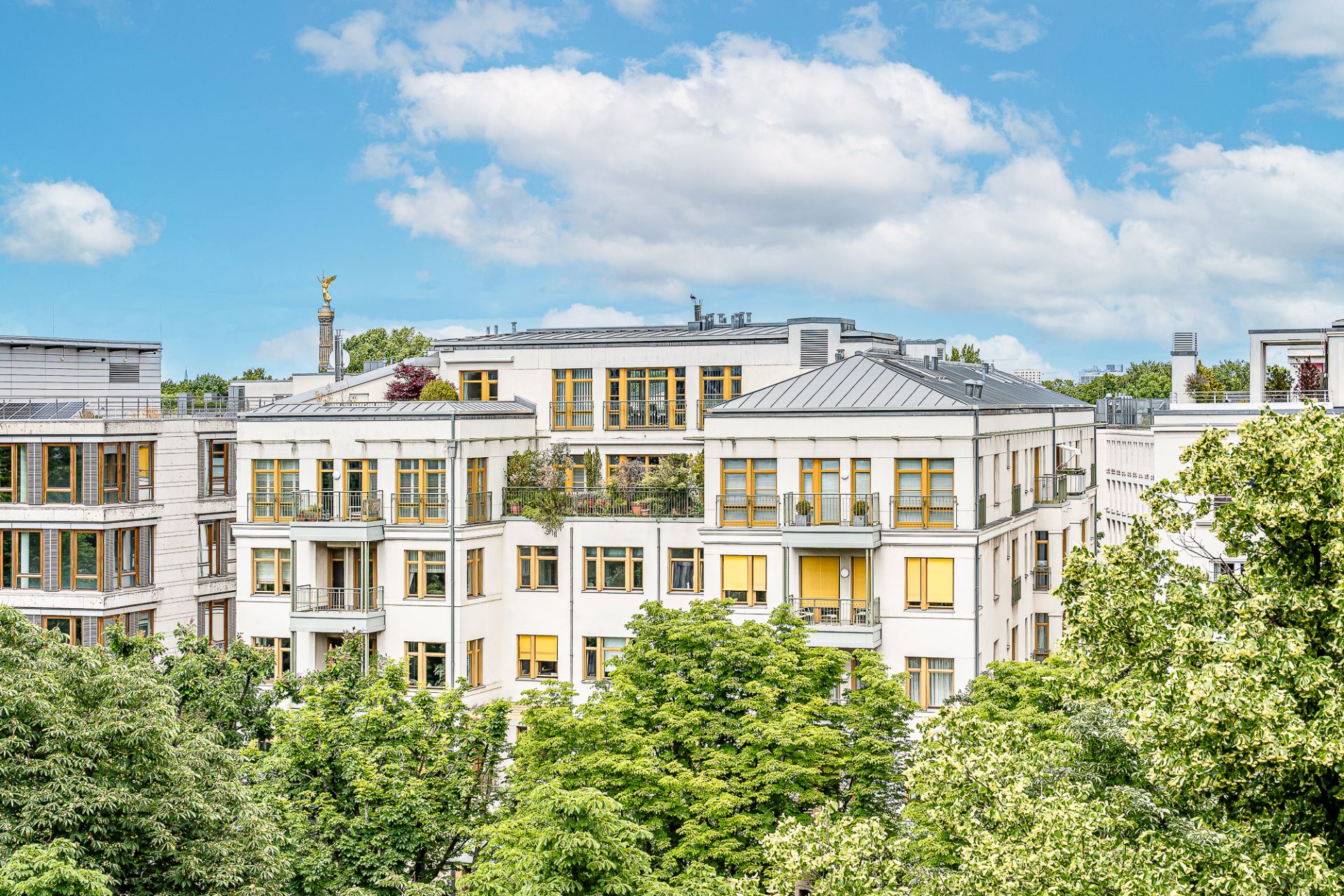 Helles Wohngebäude mit gelben Fensterrahmen, umgeben von grünen Bäumen unter blauem Himmel.