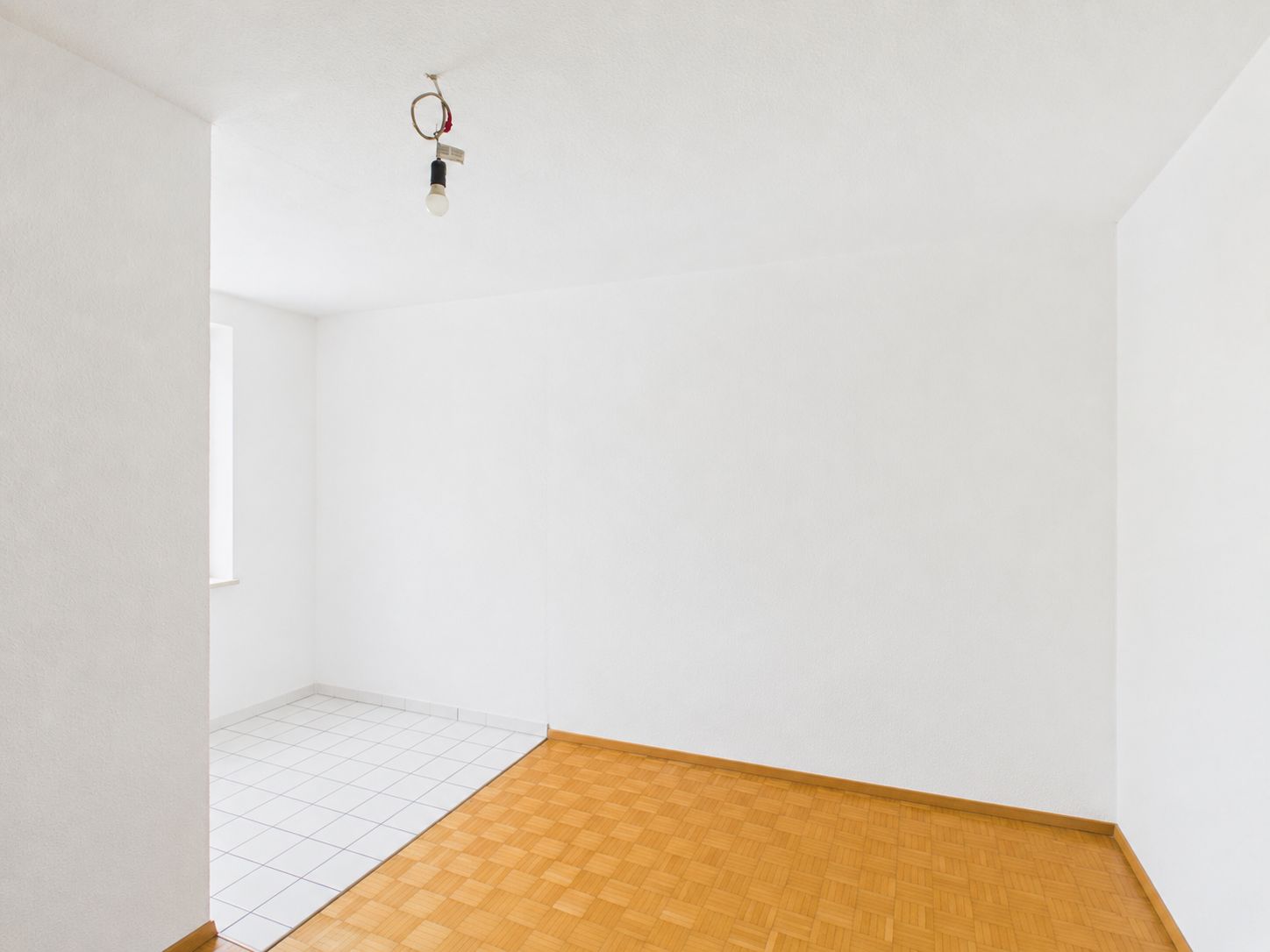 Leerer, weißer Raum mit Parkettboden und angrenzendem gefliestem Bereich, Deckenlampe ohne Abdeckung.