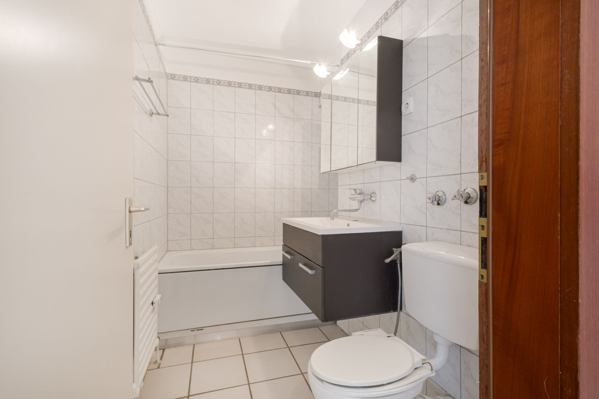 Helles Badezimmer mit Badewanne, Waschbecken, Spiegel und Toilette, weiße Fliesen an Wand und Boden.