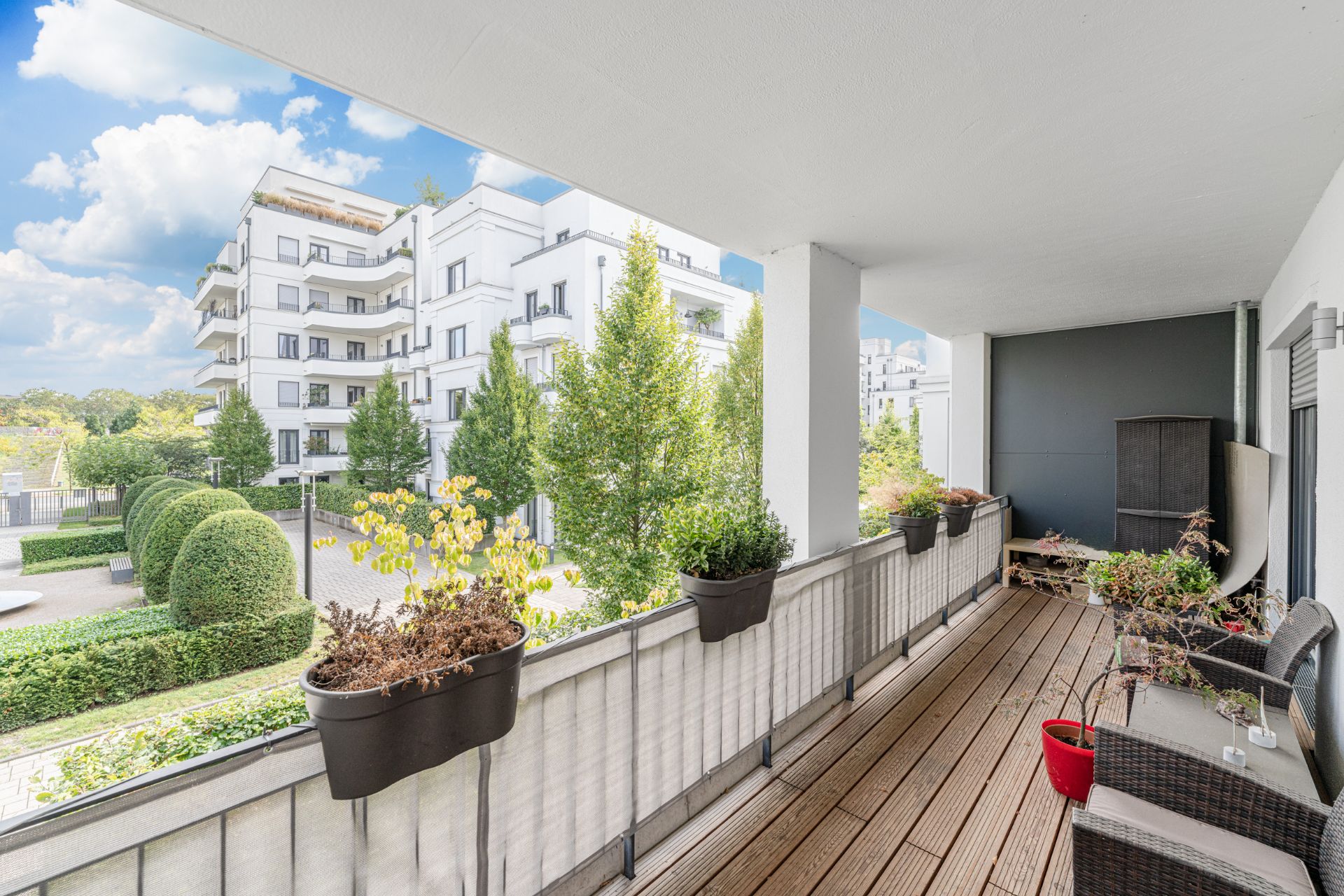 Großer Balkon mit Holzboden, Blumenkästen und Blick auf moderne Wohngebäude und Garten.