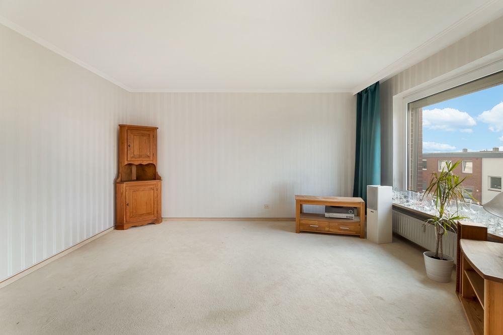 Helles Wohnzimmer mit Teppichboden, Holzmöbeln und großem Fenster mit Vorhang.
