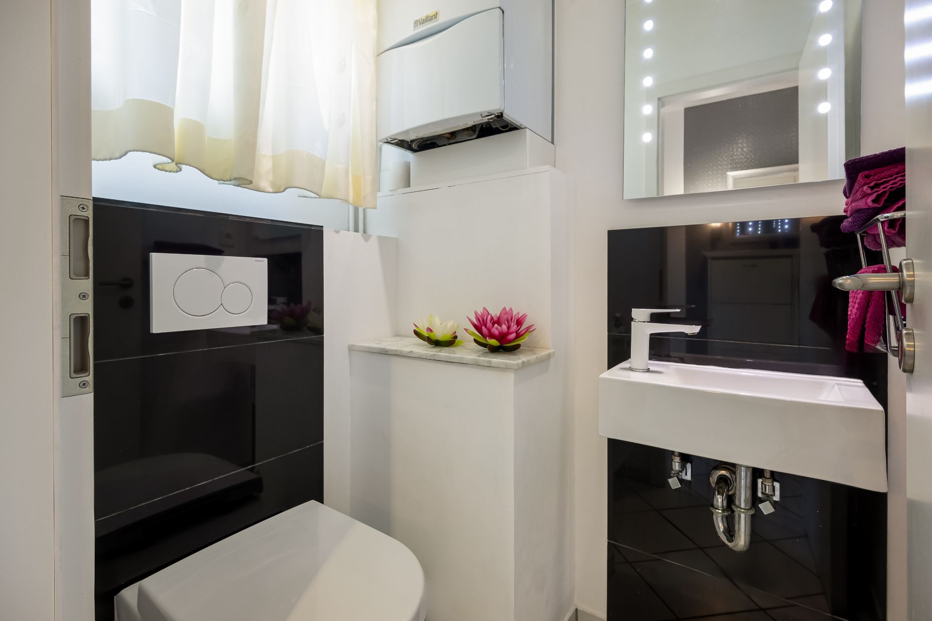 Modernes kleines Bad mit Toilette, Waschbecken, Spiegel und Dekoblumen auf einer Ablage.