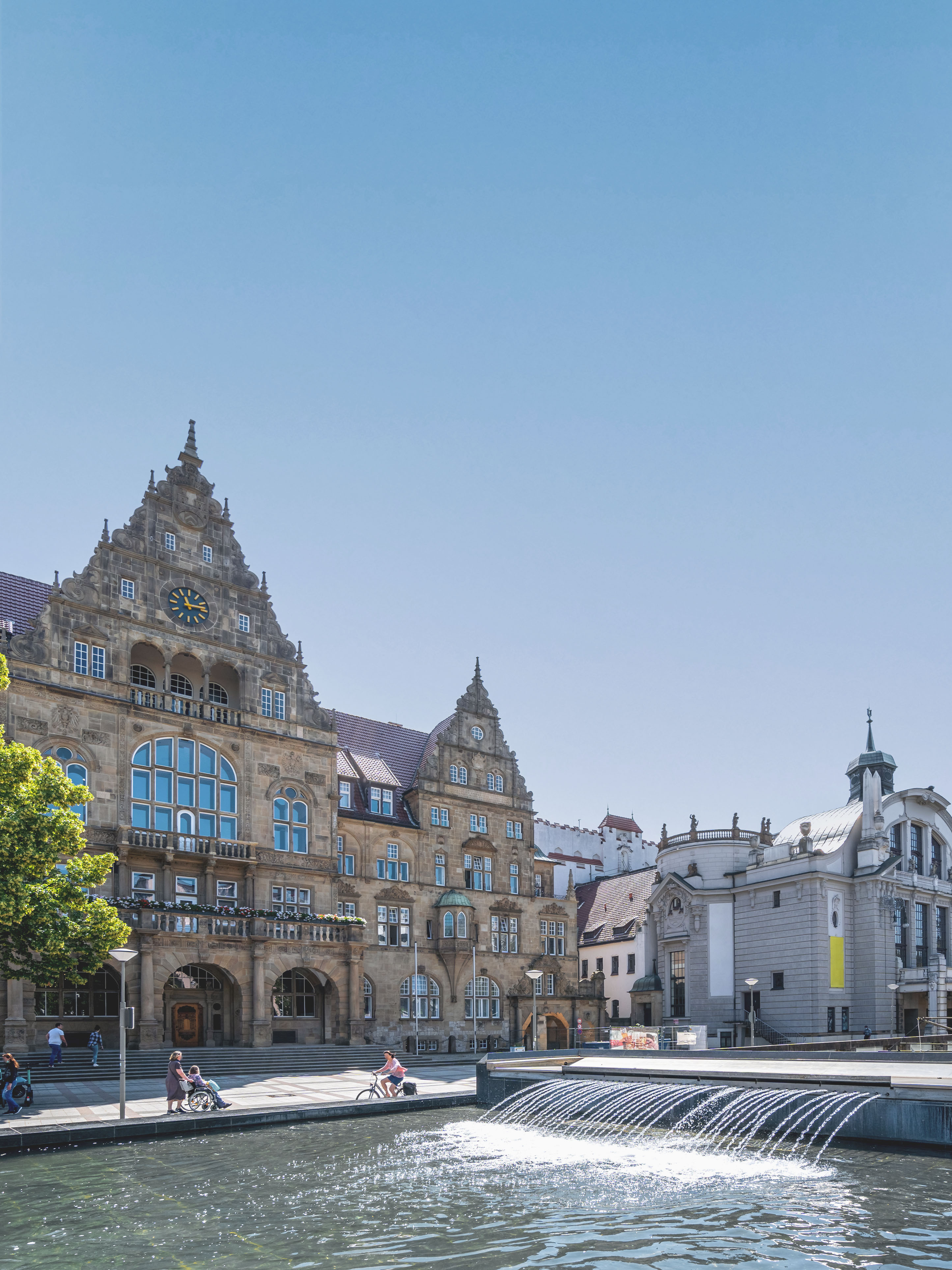 Historisches Gebäude mit Uhr und Springbrunnen davor, Menschen gehen und fahren Fahrrad.