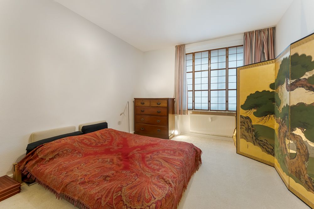 Schlafzimmer mit Doppelbett, roter Decke, Kommode, großem Fenster und bemaltem Paravent.