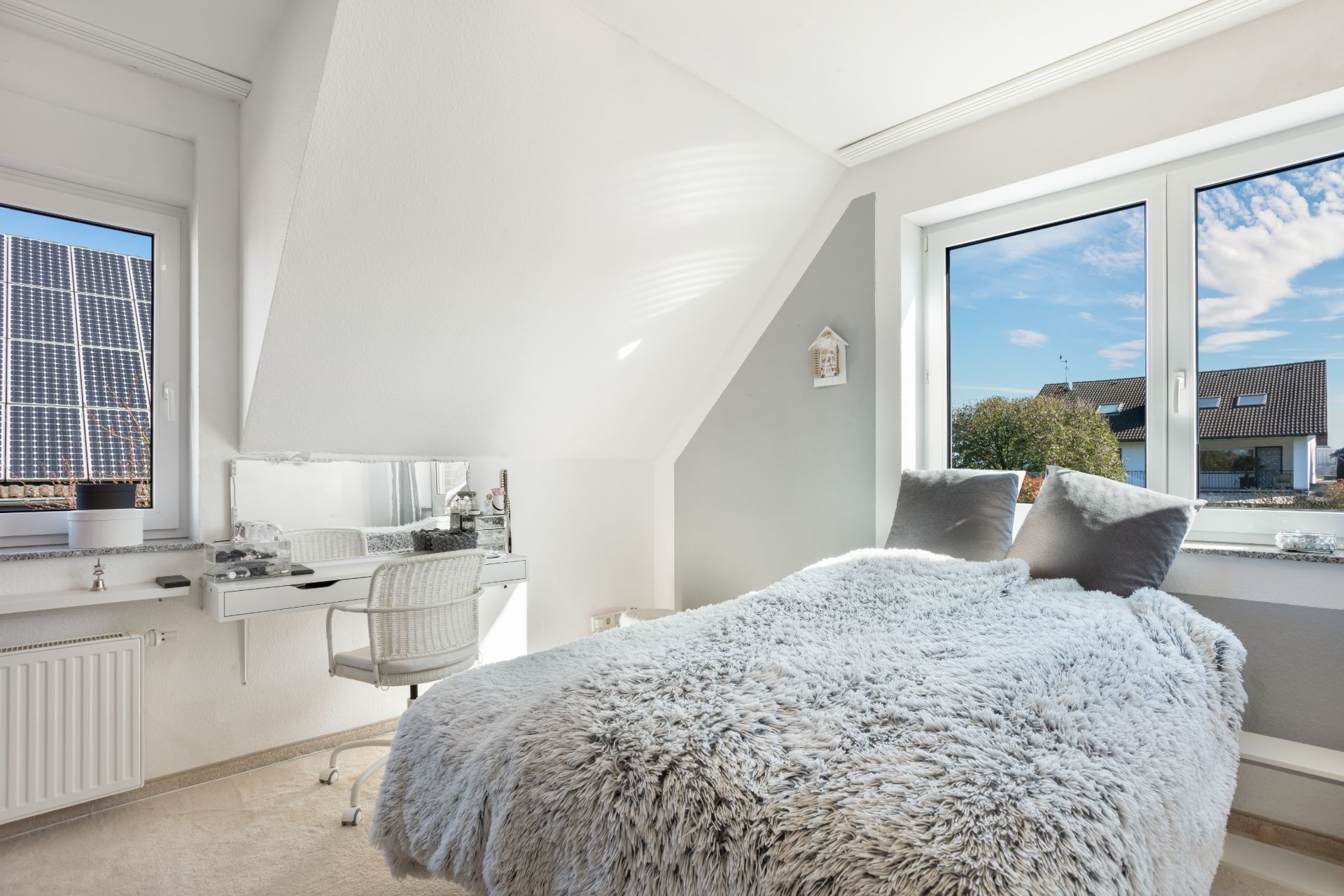 Helles Schlafzimmer mit großem Fenster, Bett mit grauer Decke und Schreibtisch am Fenster.