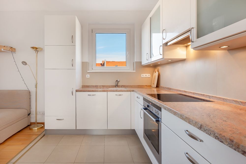 Moderne weiße Einbauküche mit Herd, Spüle und Fenster, daneben ein Sofa und Stehlampe.