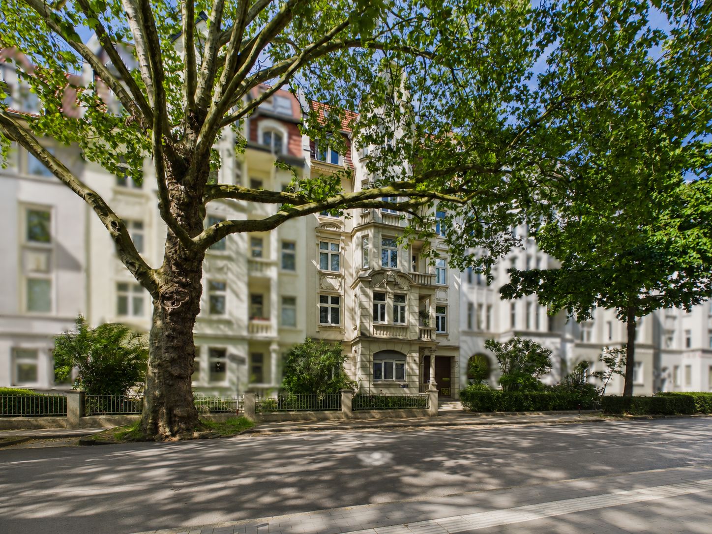 Mehrstöckiges Wohnhaus mit Vorgarten und großen Bäumen an einer ruhigen Straße.
