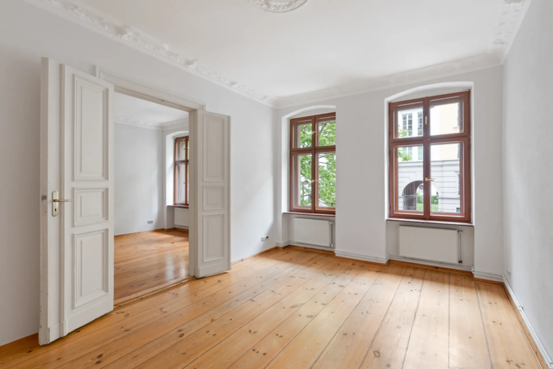 Heller, leerer Raum mit Holzboden, zwei großen Fenstern und Doppeltür zu weiterem Zimmer.