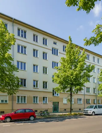Mehrstöckiges Wohnhaus mit Bäumen und Autos am Straßenrand bei sonnigem Wetter.