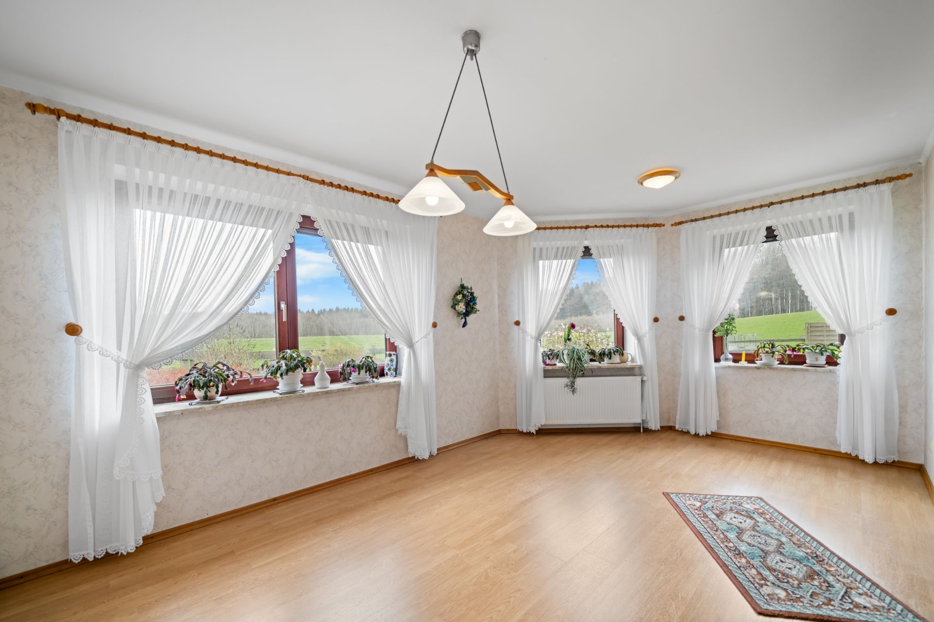 Heller Raum mit Holzfußboden, großen Fenstern, weißen Vorhängen und Pflanzen auf den Fensterbänken.
