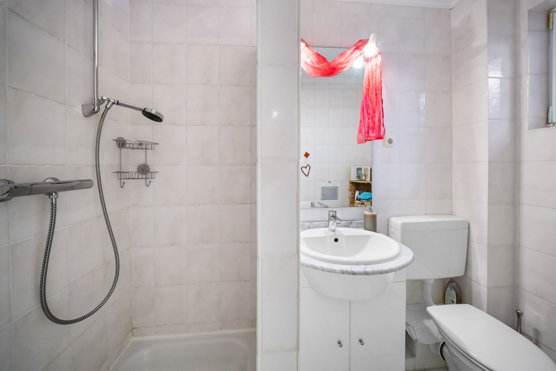 Kleines Badezimmer mit Dusche, Waschbecken, Spiegel und Toilette, weiße Fliesen an den Wänden.