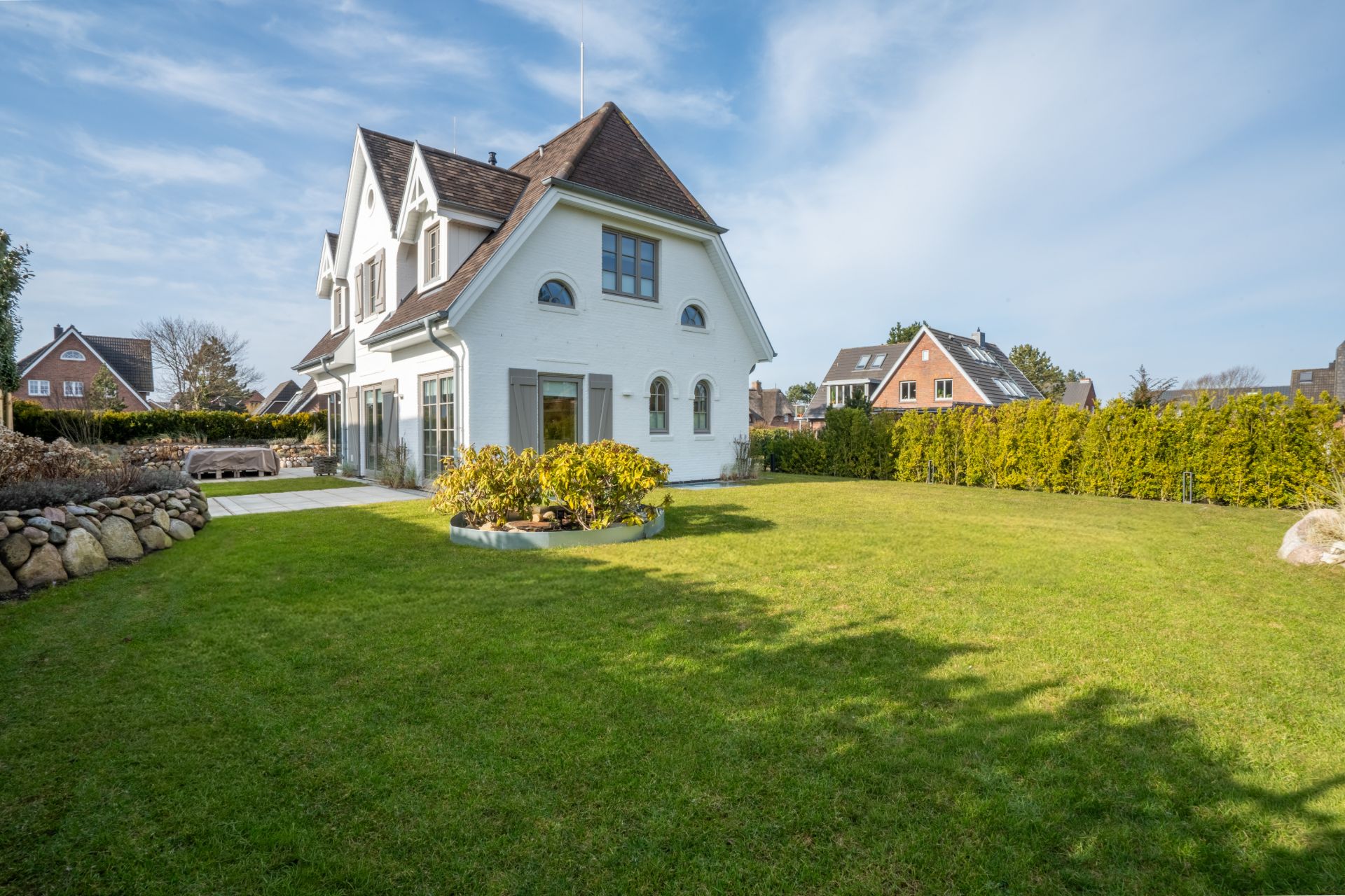 Weißes Einfamilienhaus mit großem Garten und Hecke, einige Nachbarhäuser im Hintergrund.