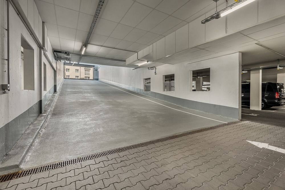 Rampe in einer Tiefgarage mit Fenstern zu Parkplätzen und einem Auto rechts.