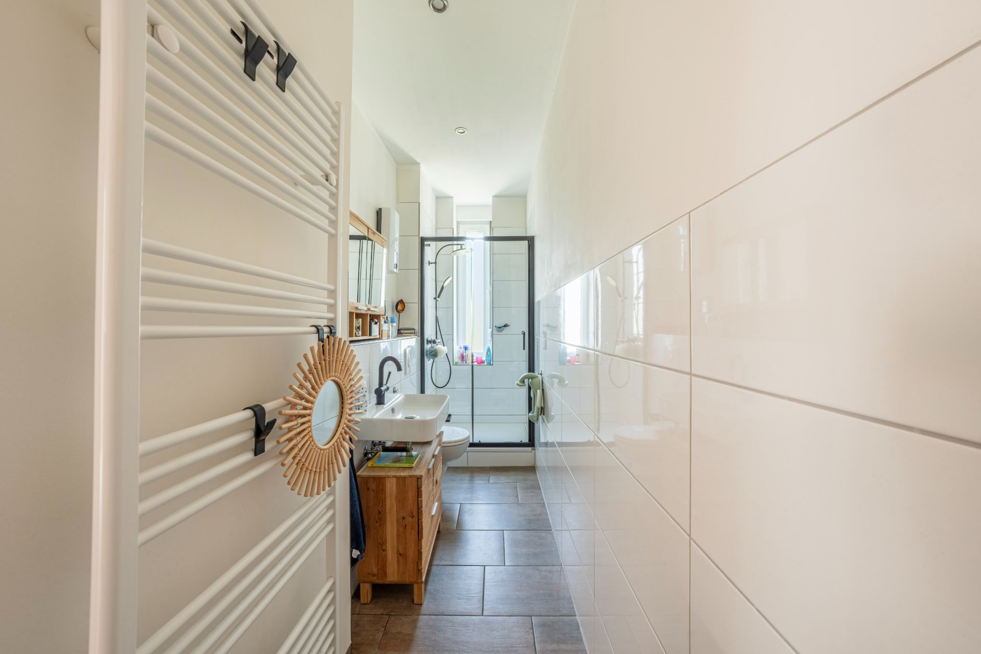 Helles Badezimmer mit Waschbecken, Spiegel, Handtuchheizung und Dusche mit Glastür.