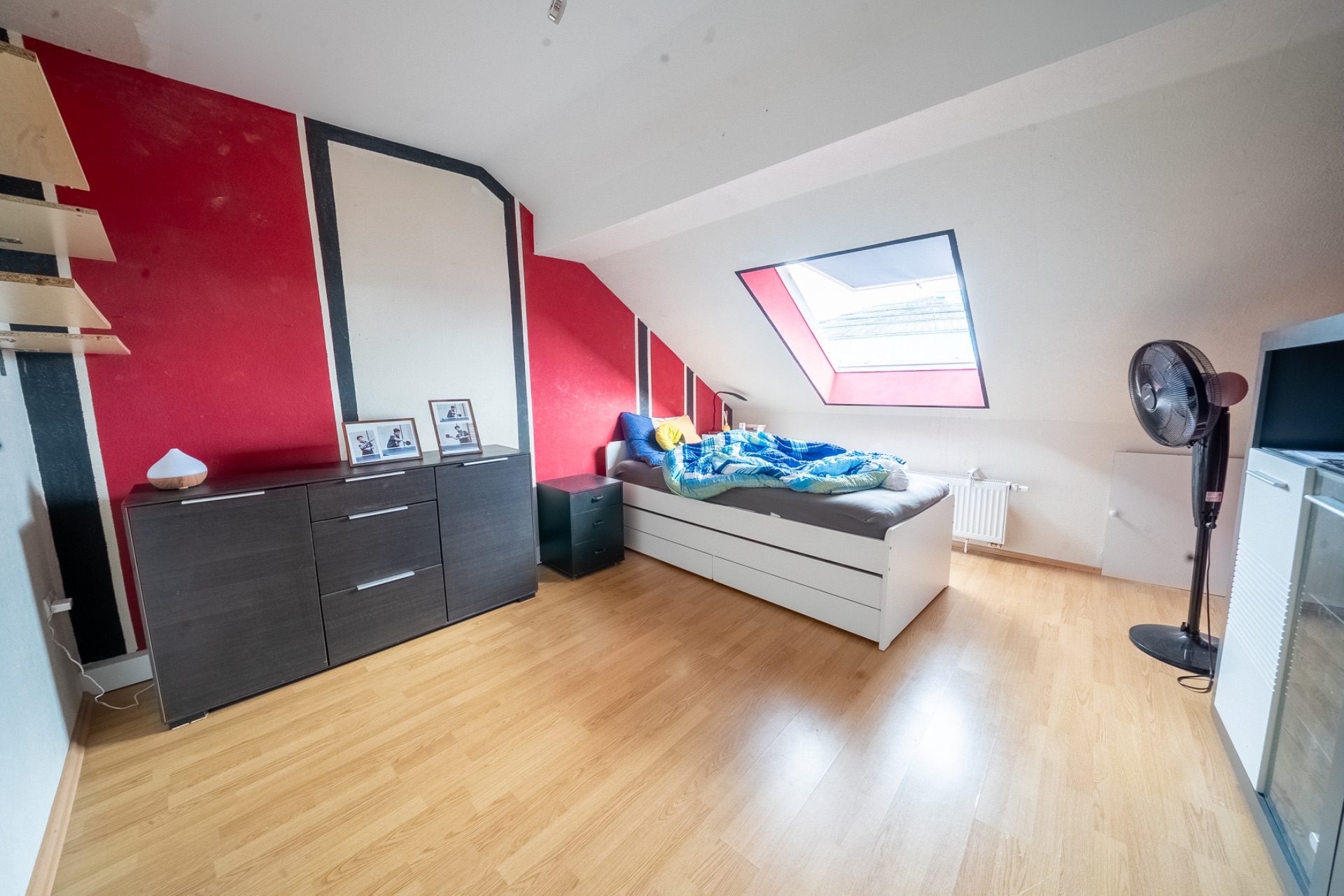 Helles Schlafzimmer mit Dachschräge, Bett, Kommode, Ventilator und großem Dachfenster.
