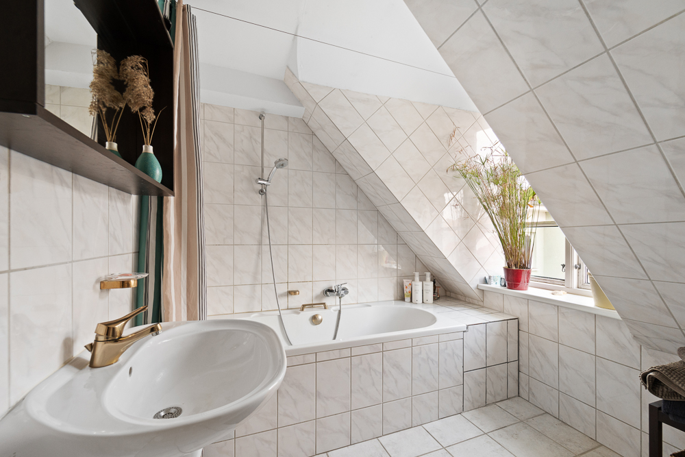 Helles Badezimmer mit Badewanne unter Dachschräge, Waschbecken und Pflanzen am Fenster.