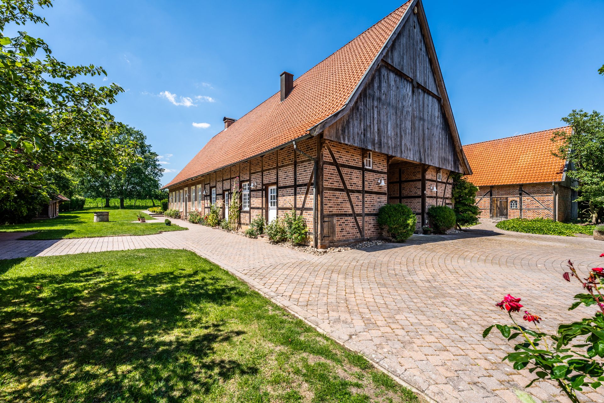 Großes Fachwerkhaus mit rotem Ziegeldach auf einem gepflasterten Hof, umgeben von grünem Garten.