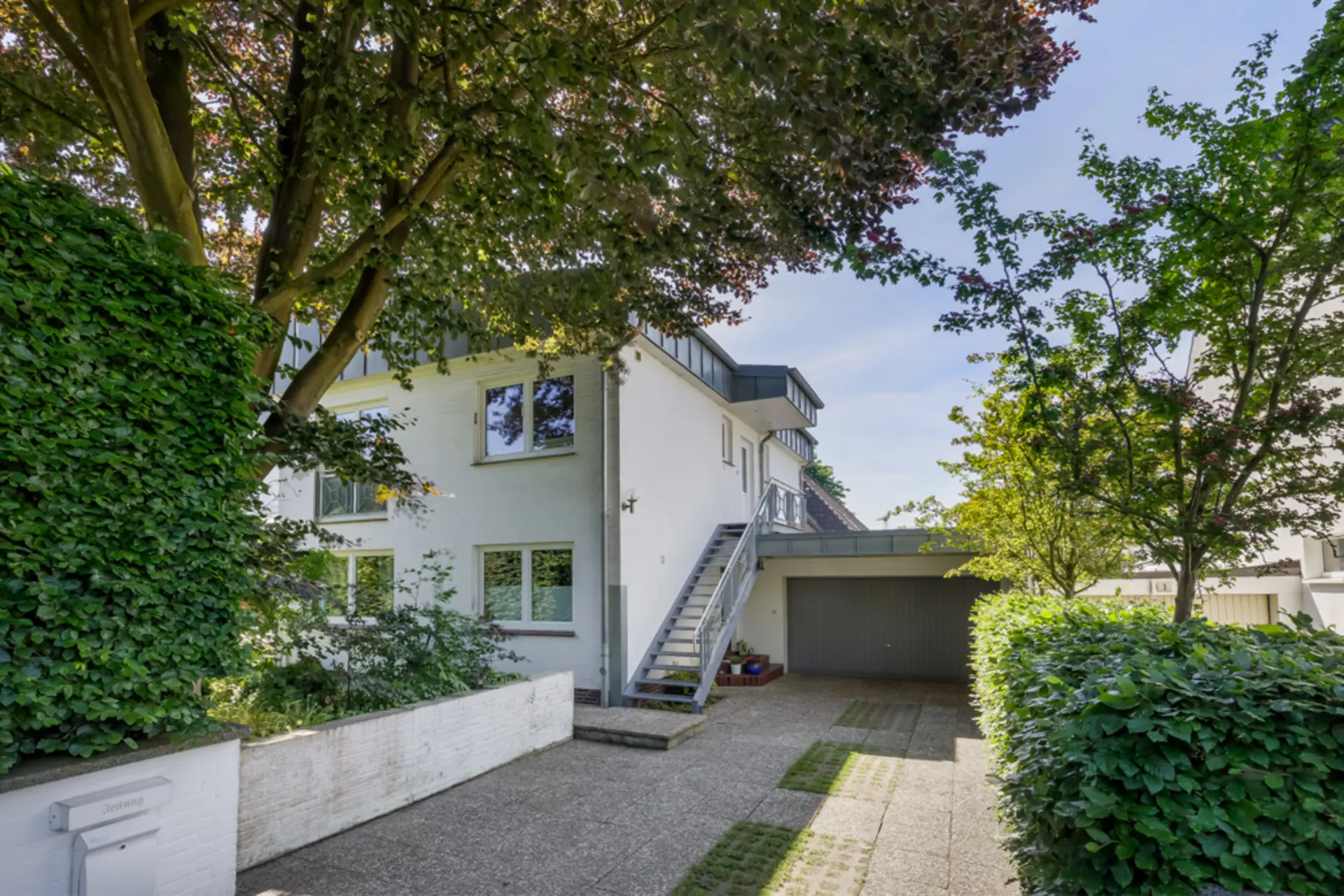 Weiße Wohnhausfassade mit Außentreppe, Garage und von Bäumen und Hecken umgebener Einfahrt.