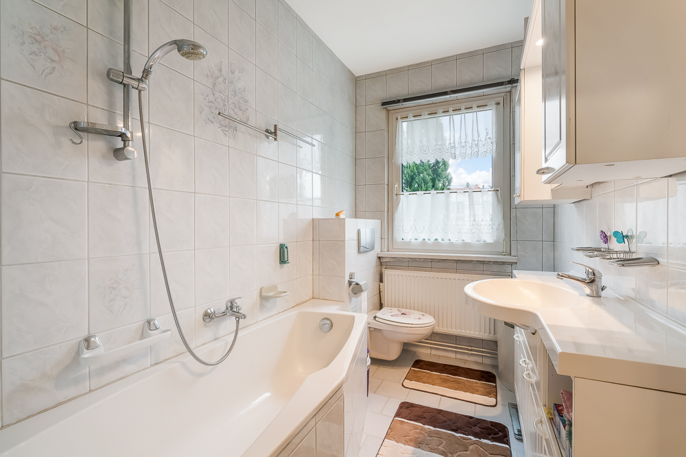Helles Badezimmer mit Badewanne, Dusche, Waschbecken, WC und Fenster mit Gardine.