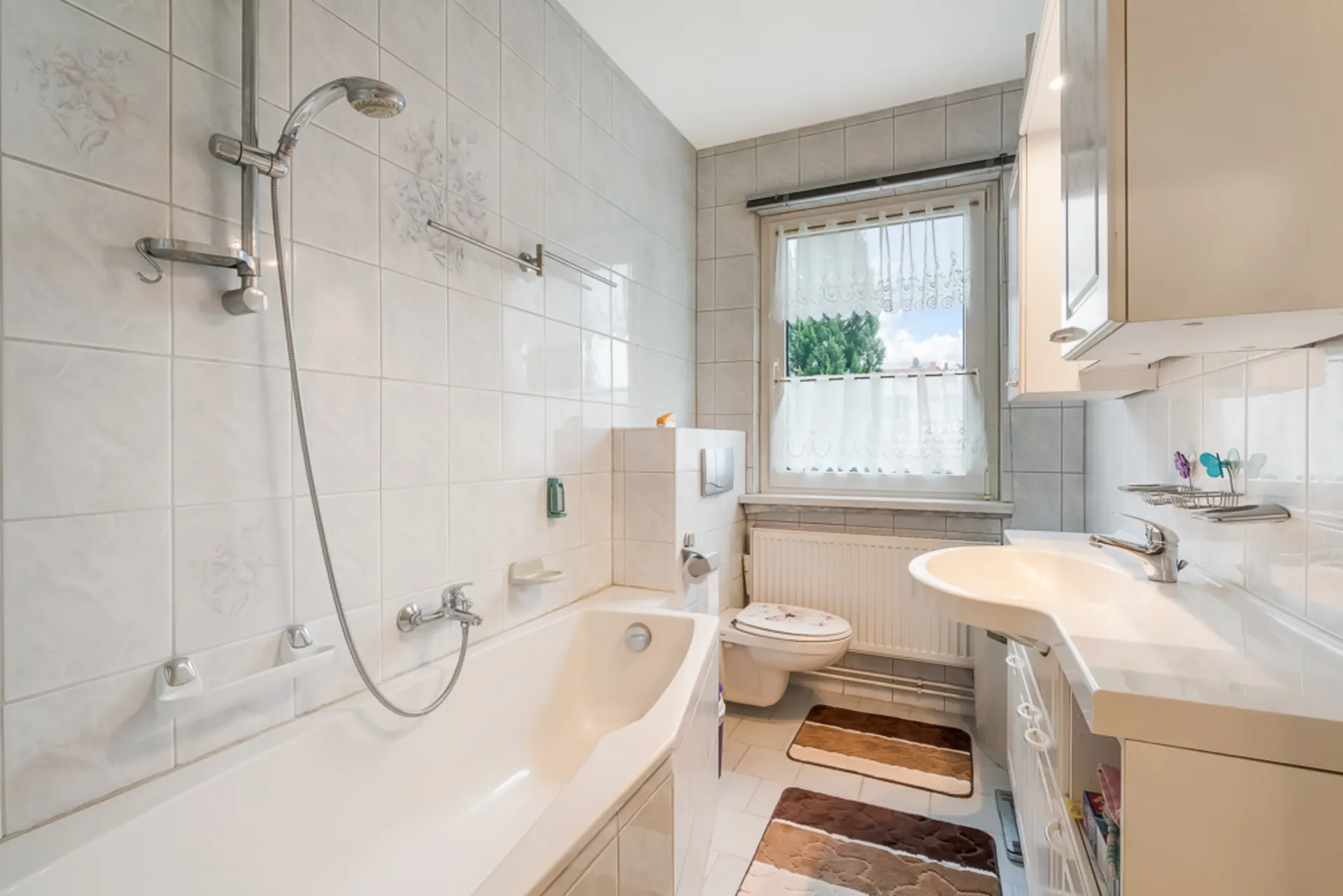 Helles Badezimmer mit Badewanne, Dusche, Waschbecken, WC und Fenster mit Gardine.